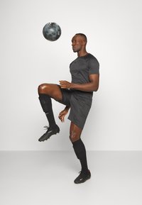 Jersey atlético negro y pantalones cortos, tejido texturizado, con un balón de fútbol negro en el aire. Medias y botas negras completan el atuendo deportivo.