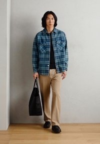 Blå rutete denimjakke over en svart skjorte, beige bukser og svarte sko. Holder en svart totebag. Enkelt design, avslappet passform.