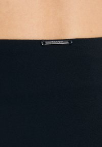 Melna auduma ar gludu tekstūru, malā ir sudraba aparatūras etiķete ar uzrakstu "Calvin Klein". Vienkāršs un elegants dizains.