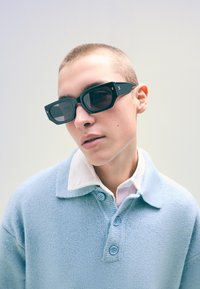 Fekete, téglalap alakú napszemüveg sötét lencsékkel és arany kiegészítőkkel, világos kék gyapjú pulóverrel és fehér galléros inggel viselve.