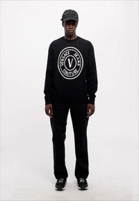Črn pleten pulover s krožno prepoznavno oznako Versace Jeans Couture, črne hlače s kontrastnimi šivi in črna kapa z oznako, združeni s črnimi športnimi copati.