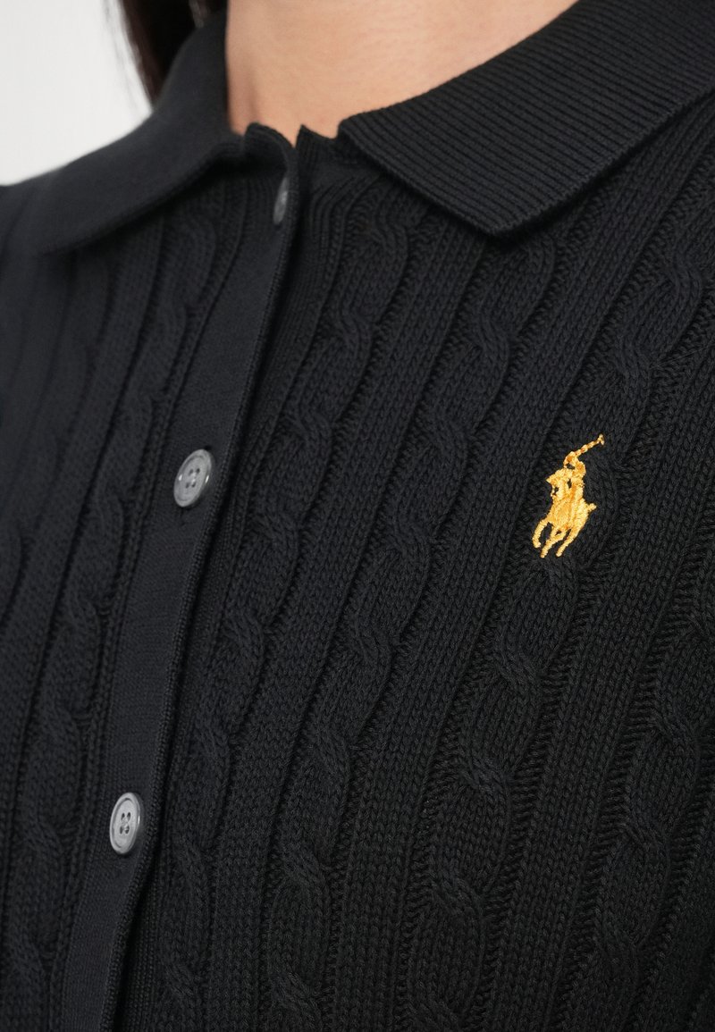 Sort kabelstrikket polobukser med ribbet krave, grå knapper og et gyldent broderet logo. Nærbillede af stoftekstur og detaljer.