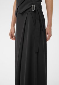 Zwarte geplisseerde rok met een zijgespannen riem en een split, gedragen door een persoon met ontspannen handen aan de zijkanten tegen een witte achtergrond.