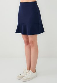 Vrouw in een donkerblauwe uitlopende rok en witte sneakers, staand voor een effen witte achtergrond.
