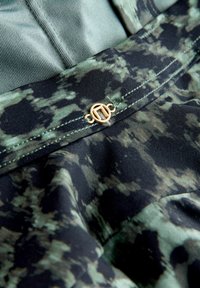 Schwarz-grün gemusteter Stoff mit glänzender Textur, versehen mit einer goldfarbenen dekorativen Schnalle am Taillenbund. Nahaufnahme der Nähdetails.