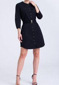 Svart knäppt klänning i mjukt tyg, med lång ärm, två bröstfickor, elastisk midja med ett guldspänne, och en utsvängd nederdel.