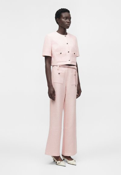 Femme debout portant une veste texturée rose pâle à manches courtes et un pantalon large assorti, associée à des talons blancs à bouts pointus.