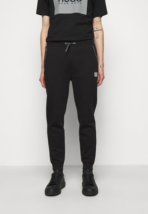 Zwarte joggingbroek met een trekkoord in de taille, zijzakken met rits en een grijze logopatch. Gecombineerd met een zwart T-shirt met een grijze afbeelding.