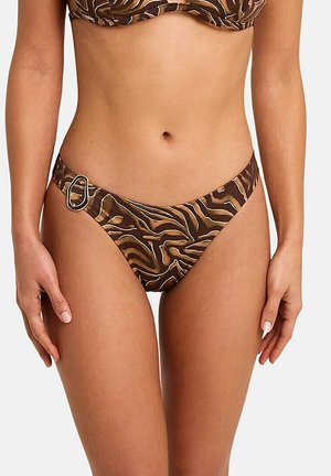 Femme portant un bas de bikini brun à motif zébré avec une boucle métallique décorative sur la hanche gauche, sur un fond uni.