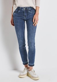 Wąskie niebieskie dżinsy z przetarciem, srebrnym zapięciem i postrzępionym dołem, zestawione z białymi i beżowymi sneakerami.