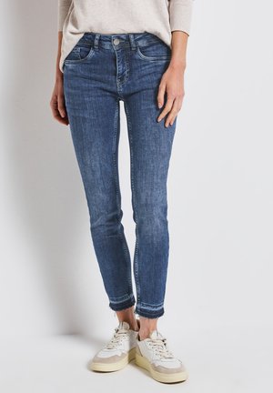 Jeans de mezclilla ajustados de lavado desgastado, con cierre de botón plateado y dobladillo deshilachado, combinados con zapatillas blancas y beige.