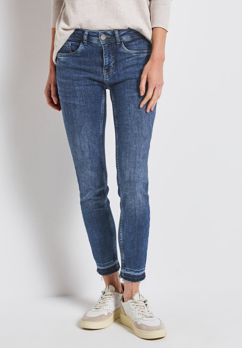 Wąskie niebieskie dżinsy z przetarciem, srebrnym zapięciem i postrzępionym dołem, zestawione z białymi i beżowymi sneakerami.