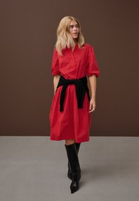 Femeie blondă purtând o rochie roșie până la genunchi cu mâneci bufante, un pulover negru legat în talie și cizme negre până la genunchi.