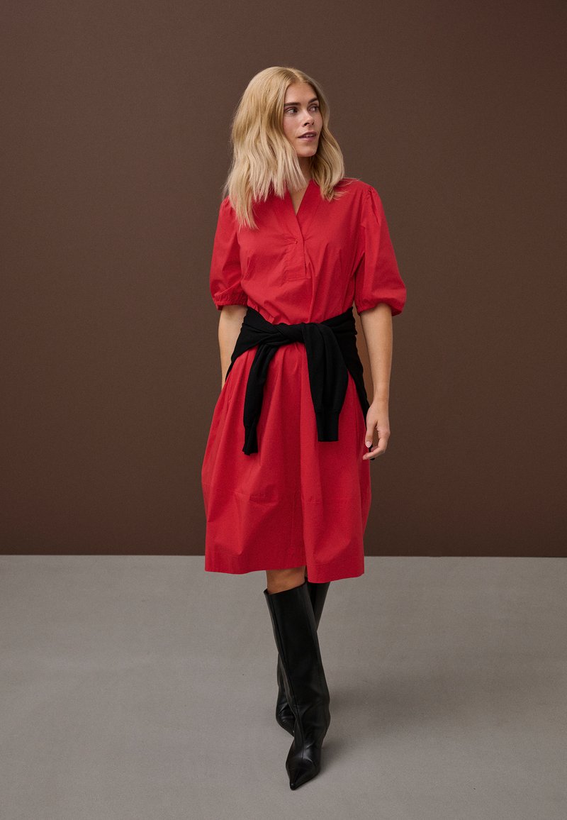 Femeie blondă purtând o rochie roșie până la genunchi cu mâneci bufante, un pulover negru legat în talie și cizme negre până la genunchi.