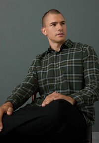 Chemise à carreaux verts en tissu avec un devant boutonné, posée sur un fond atténué, portée avec un pantalon noir, en posture assise.