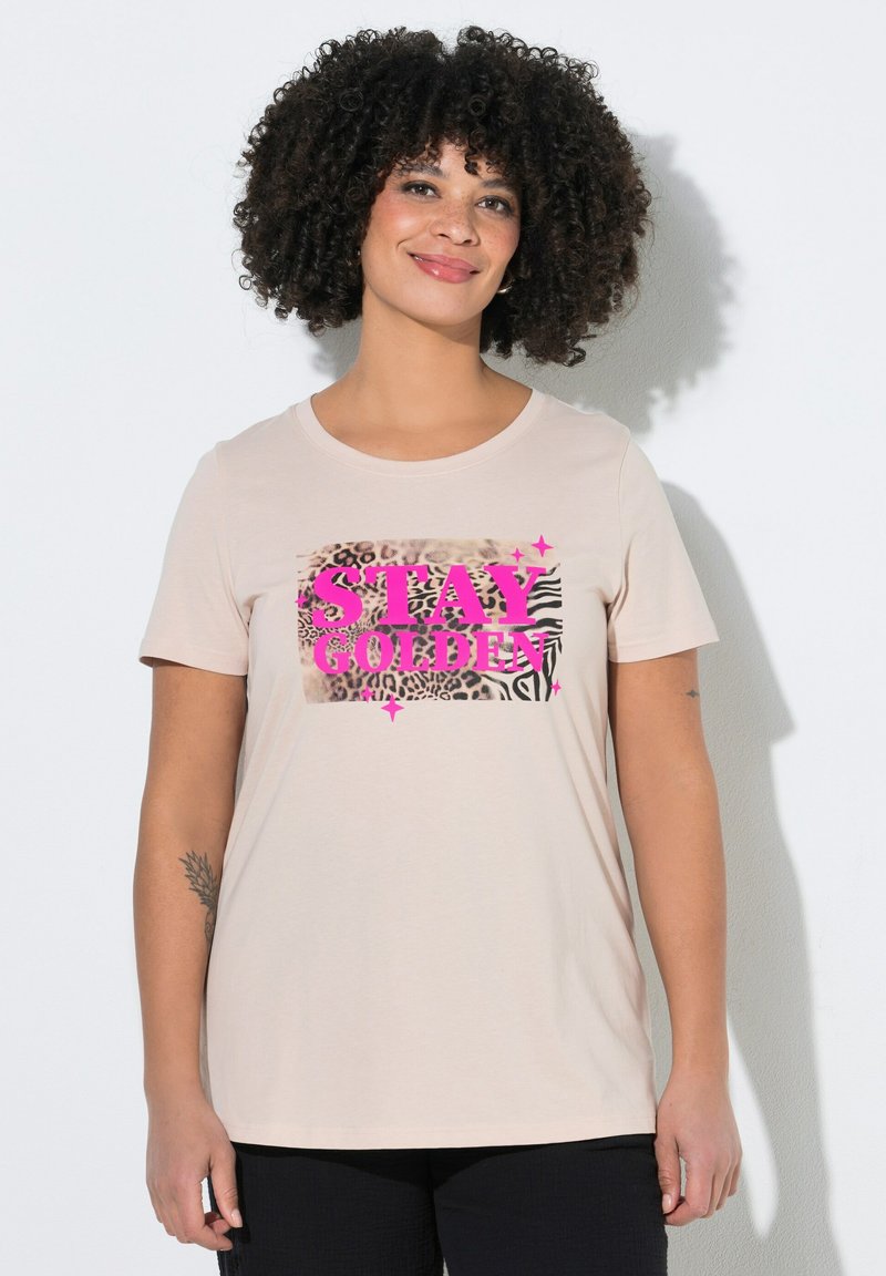 Femme aux cheveux bouclés portant un t-shirt beige clair avec un motif animalier en bloc et un texte rose vif "STAY GOLDEN", debout contre un mur blanc.