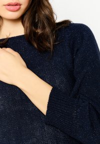 Navy blue knitted sweater with a textured finish, featuring long sleeves and a loose fit. The detail showcases a slightly ribbed cuff.