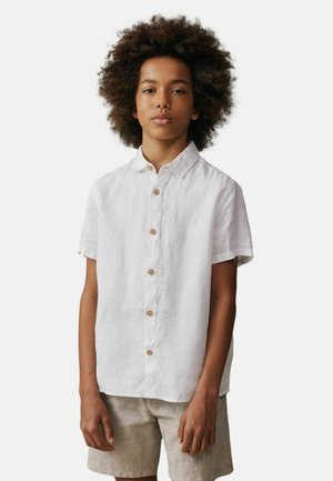 Jongen met krullend haar, gekleed in een wit kortmouwig overhemd met knopen en beige korte broek, staand tegen een effen achtergrond.