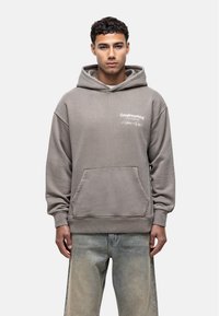 Haut à capuche en coton gris avec une poche kangourou, des poignets et un ourlet côtelés. Présente un motif imprimé blanc à l'avant. Porté avec un jean bleu délavé.