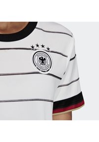 Maglia sportiva bianca con strisce orizzontali nere, colletto nero, emblema della Federazione Calcistica Tedesca, e rifiniture nelle maniche rosse, nere e dorate su una persona.