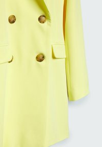 Dubbelgeknoopte jas van felgele stof, met goudkleurige knopen en twee zakken aan de voorkant, met een rechte snit en lange mouwen.