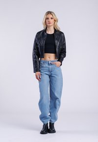 Černá zkrácená kožená bunda přes černý top, v kombinaci s volnými světle modrými džínami a černými kotníkovými botami, na neutrálním pozadí.