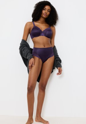 Fioletowy satynowy biustonosz z koronkowymi akcentami, zestawiony z dopasowanymi figami z wysokim stanem. Modelka ma na sobie szary kardigan. Gładka faktura i delikatne detale.