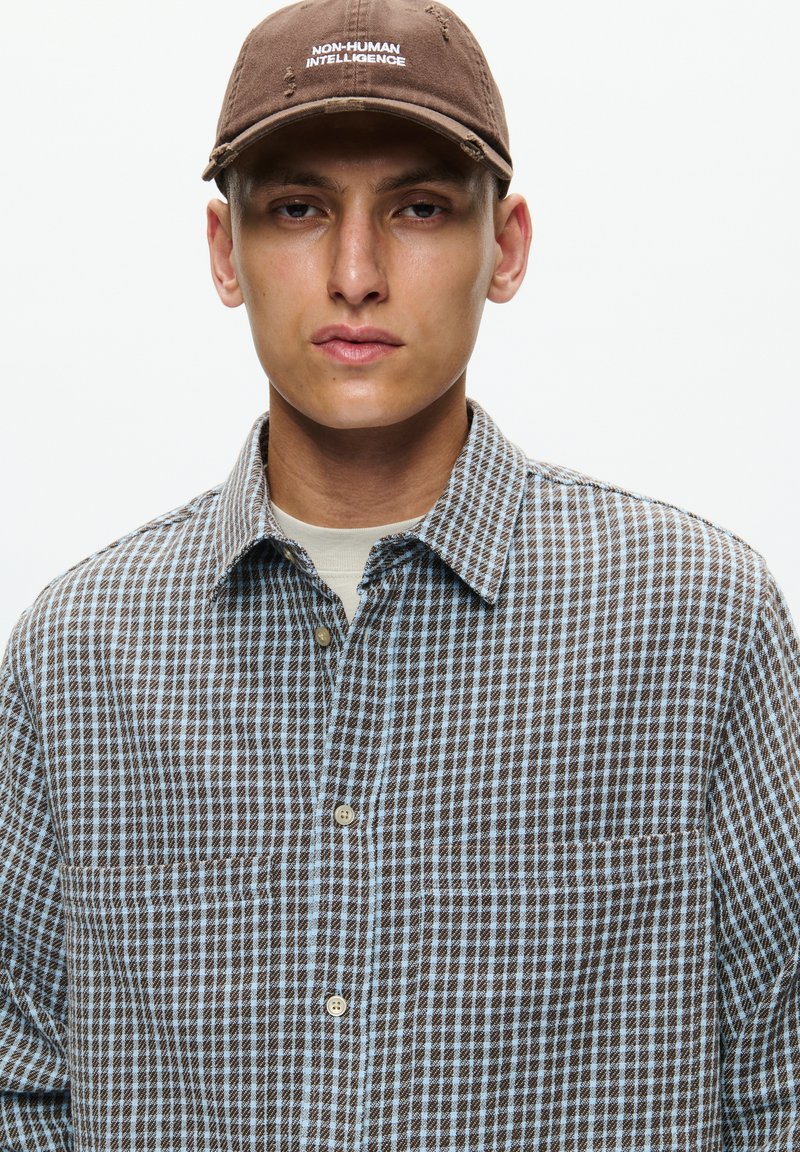 Young man wearing a brown cap with "NON-HUMAN INTELLIGENCE" text and a blue and brown checkered button-up shirt.
