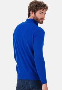 Pull bleu tricoté avec un col haut, coupe ajustée, poignets et ourlet à côtes, texture lisse. Porté avec un pantalon en denim foncé.