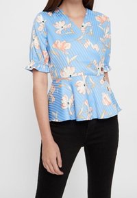 Top peplum floreale blu con scollo a V e maniche corte arruffate, decorato con fiori rosa e crema su uno sfondo a righe. Abbinato con pantaloni neri.