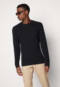 Black textured knit sweater with a crew neckline and ribbed cuffs, paired with beige pants. Sweater features a fitted silhouette.