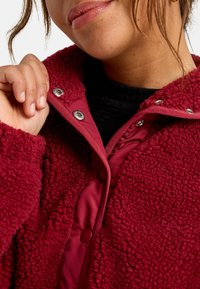 Giacca rossa texturizzata con esterno in pile e interno liscio, dotata di chiusure a scatto e colletto alto, che mostra parte di una maglietta nera sottostante.