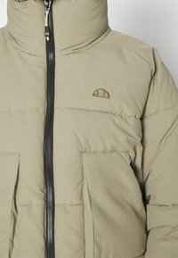 Light olive-green puffer jacket with a high collar, front zipper, and large front pockets. Embroidered logo on the chest. Smooth texture.