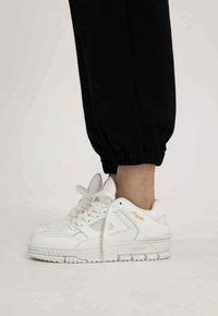 Witte leren sneakers met een gestructureerd ontwerp, ronde neus en prominente gouden "ARIGATO" accent aan de zijkant. Gecombineerd met zwarte opgerolde broek.