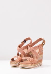 Une paire de sandales compensées en toile beige avec des brides entrecroisées et une boucle à la cheville, posées sur une surface blanche avec un fond uni.