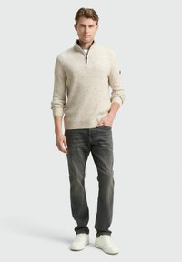 Jonge man staat met één hand in zijn zak, gekleed in een beige rits-trui, donkergrijze spijkerbroek en witte sneakers, tegen een lichte achtergrond.