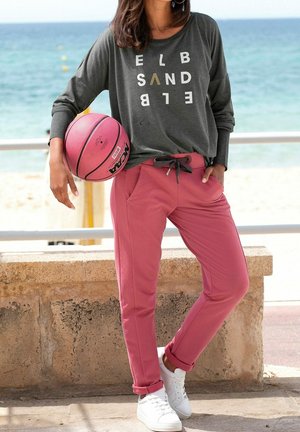 Vrouw staat bij de reling van het strand en houdt een roze basketbal vast, draagt een grijs shirt met lange mouwen met tekst en roze broek met trekkoord, en witte sneakers.
