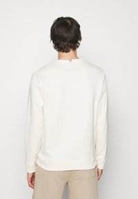 Light cream sweatshirt with long sleeves, crew neckline, and ribbed cuffs. Features small tri-color accents on the back. Soft texture.