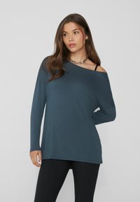 Langärmliges, schulterfreies Oberteil aus blau-grauem Stoff. Weiche Textur mit lockerer Passform. Kombiniert mit schwarzen Leggings und einer Kettenhalskette.