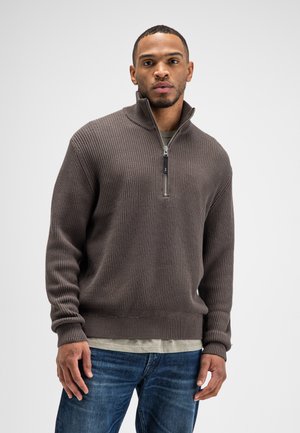 Man draagt een bruin geribd half-zip vest over een beige overhemd en blauwe spijkerbroek, staand voor een effen lichtgrijze achtergrond.