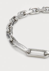 Ez a sima felületű, összekapcsolódó láncszemekből álló ezüst lánc karkötő egy "Calvin Klein" felirattal ellátott címkét is tartalmaz.