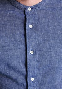 Nærbillede af en person iført en blå chambray skjorte med knapper og stående krave samt hvide knapper.