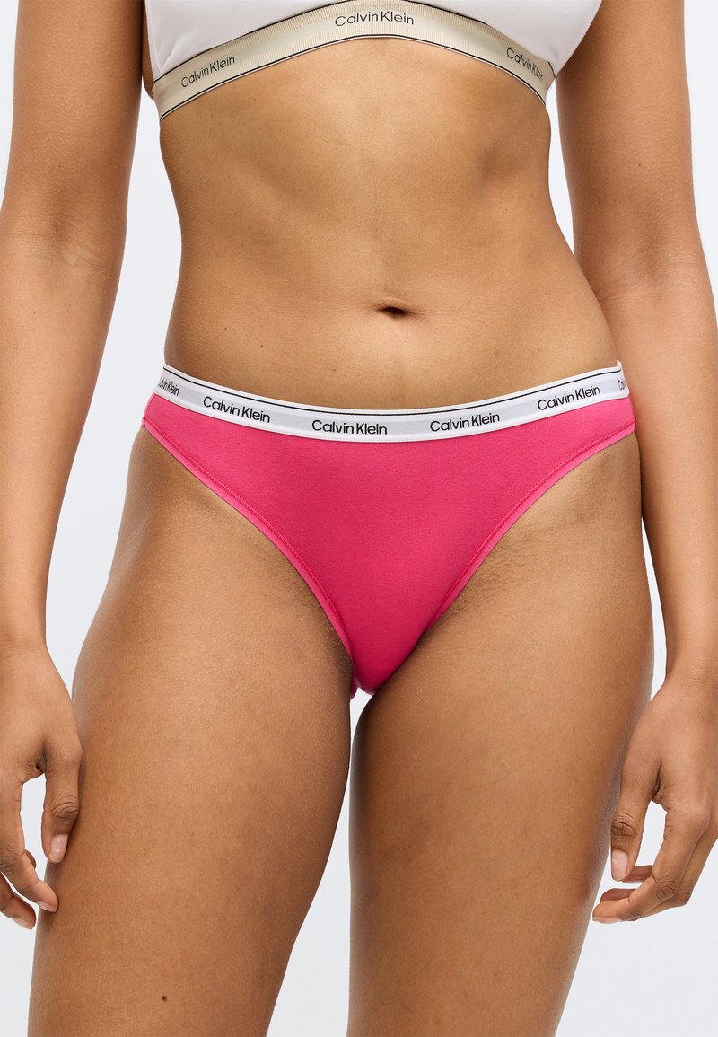 Sieviete, tērpusies rozā Calvin Klein bikini apakšveļā un baltā Calvin Klein sporta krūšturī, stāv ar rokām atviegloti pie sāniem.