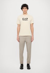 Béžové bavlněné tričko s černě potištěným logem "EA7 EMPORIO ARMANI." V páru se světle šedými kalhotami a černými botami.