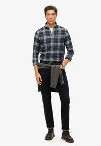 Camicia a quadri blu, verde e bianco; pantaloni scuri; maglione grigio annodato in vita; indossa stivali neri e un orologio da polso.