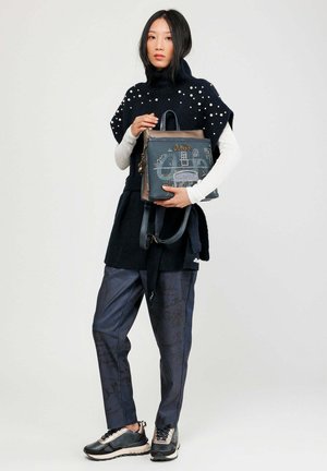 Blaues Handtasche mit strukturiertem Muster, versehen mit braunen Akzenten und einem Reißverschluss, gehalten von einem Modell in einem schwarz-weißen Outfit.