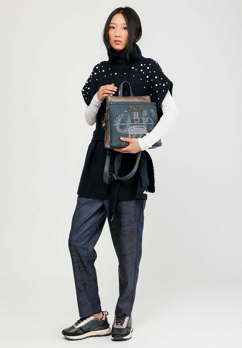 Sac à main bleu avec un motif texturé, doté d'accents marron et d'une fermeture éclair, tenu par un mannequin vêtu d'une tenue en noir et blanc.