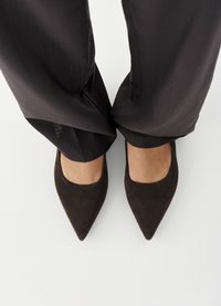 Πόδια που φορούν σκούρα καφέ φλατ παπούτσια με μυτερή μύτη, συνδυασμένα με μαύρο παντελόνι με φαρδιά μπατζάκια, σε λευκό πάτωμα.