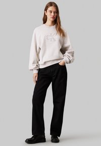 Sudadera gris claro con el logo "CK" bordado, corte relajado y mangas acampanadas, combinada con pantalones de pierna ancha negros y zapatos negros.