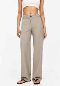 Pantalon large beige au toucher lisse, avec une fermeture à bouton, des passants de ceinture et une coupe ajustée. Associé à des tongs noires.